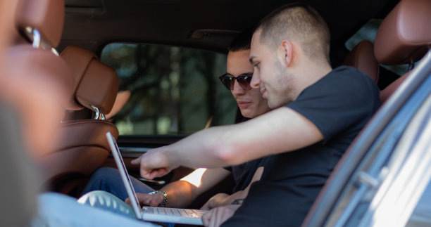 两个朋友坐在车的后座上，看着笔记本电脑，为他们的公路旅行制定计划，寻找他们的目的地。高清库存图片。图片下载