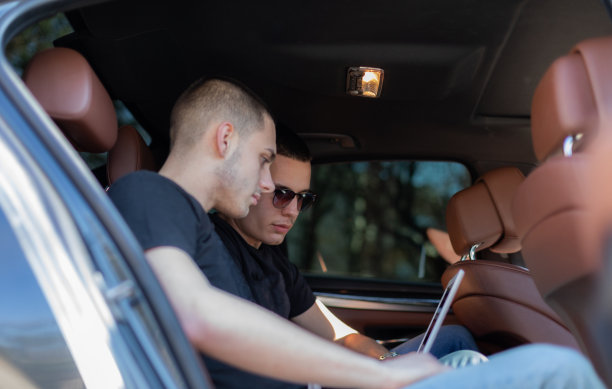 两个朋友坐在车的后座上，看着笔记本电脑，为他们的公路旅行制定计划，寻找他们的目的地。高清库存图片。图片下载