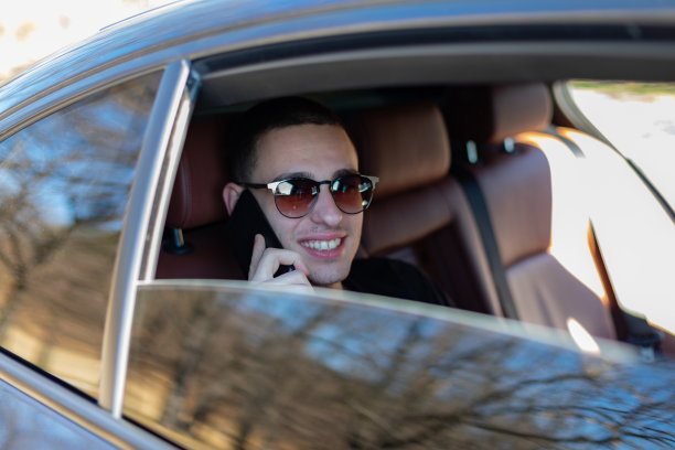 高清照片，一个年轻人坐在汽车的后座上用自己的手机在路上，制定计划，搜索，上网和媒体，聊天，微笑，享受公路旅行在一个美丽的阳光明媚的日子。图片下载