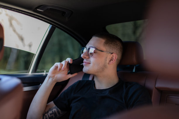 高清照片，一个年轻人坐在汽车的后座上用自己的手机在路上，制定计划，搜索，上网和媒体，聊天，微笑，享受公路旅行在一个美丽的阳光明媚的日子。图片下载