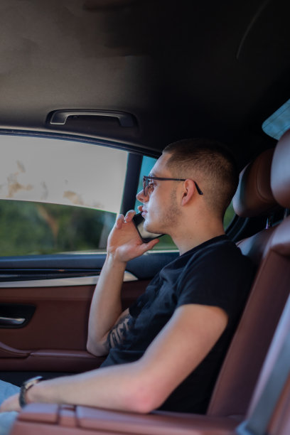 高清照片，一个年轻人坐在汽车的后座上用自己的手机在路上，制定计划，搜索，上网和媒体，聊天，微笑，享受公路旅行在一个美丽的阳光明媚的日子。图片下载