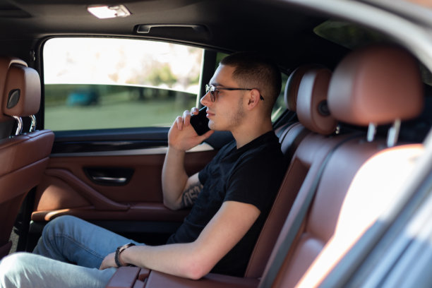 高清照片，一个年轻人坐在汽车的后座上用自己的手机在路上，制定计划，搜索，上网和媒体，聊天，微笑，享受公路旅行在一个美丽的阳光明媚的日子。图片下载