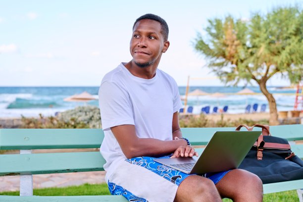 年轻微笑的自由职业者男性在沙滩上放松，坐在长凳上使用笔记本电脑图片下载