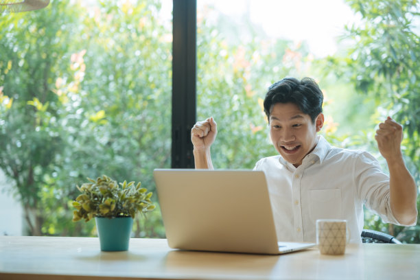 亚洲男子在家里用笔记本电脑工作时庆祝成功。图片下载