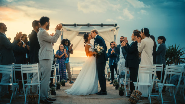 美丽的新娘和新郎庆祝婚礼在户外海滩附近的海洋日落。完美的婚礼场所，最好的来自不同民族的朋友向新婚夫妇扔花瓣。图片下载