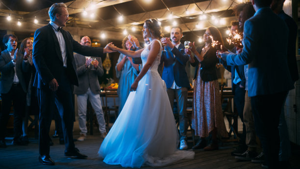 美丽的新娘在白色礼服和新郎在时尚的黑色西装庆祝婚礼在一个晚上的接待派对。新婚夫妇与来自不同民族的朋友在一起跳舞。图片下载