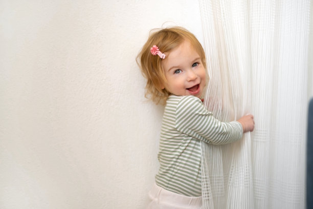 1、2岁的可爱的白种人金发女孩玩白色窗帘，在家里的灯光室内看着相机玩得很开心。孩子，孩子，幼儿在室内玩游戏，大笑，微笑的婴儿。本空间图片下载