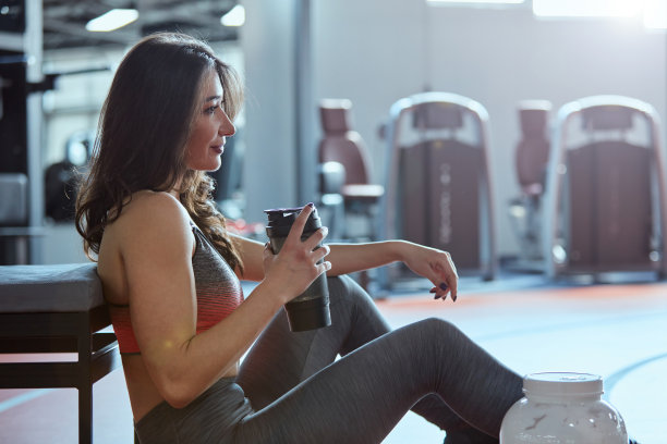 年轻健康的女人坐在健身房的地板上，放松着，喝着蛋白质奶昔。图片下载