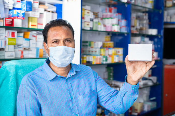 药剂师带着医用口罩在药店看着摄像头展示药盒——covid-19冠状病毒药物的概念、产品推荐和广告。图片下载