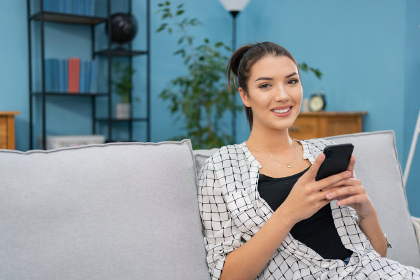 年轻女子坐在客厅的沙发上，手里拿着手机，女孩看着智能手机上的图片，微笑，和朋友聊天，上网，在屏幕前度过她的空闲时间图片下载