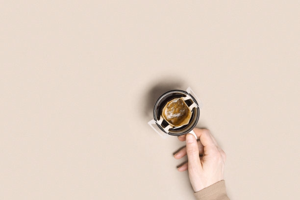 女人手握杯子，使黑咖啡喝在杯子，滴咖啡袋与研磨咖啡煮在杯子，纸袋滴咖啡过滤器，米色背景，俯视图，最小平坦的lay图片下载