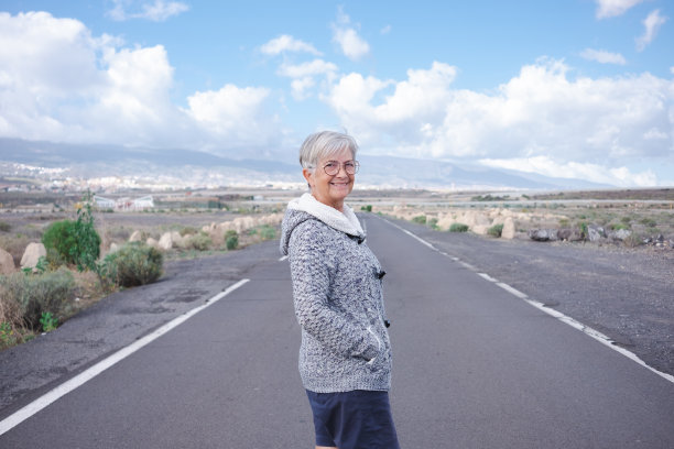 微笑独自旅行者年长妇女走在户外享受自由看着相机。空荡的道路上，苍茫的山峦和蔚蓝的天空上，有一位成熟的老妇人图片下载