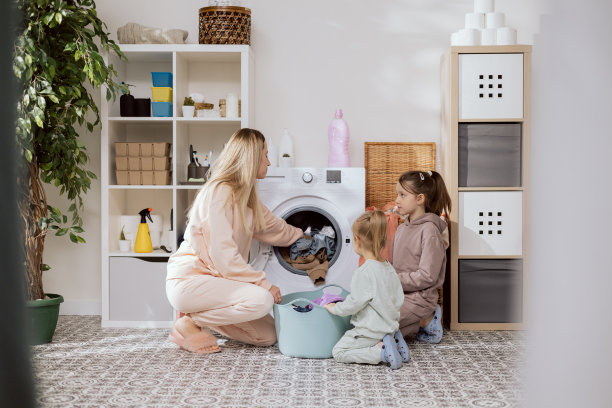照片从后面的衣架上的衣服，洗衣房，浴室的母亲和两个女儿跪在洗衣机旁边的碗与衣服妇女扔脏东西到鼓纺纱干燥图片下载