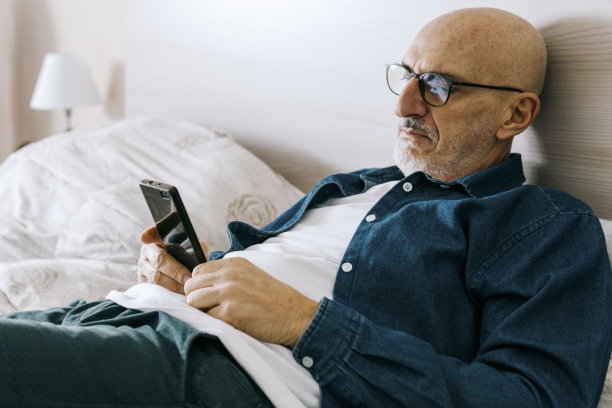 中年男子拿着智能手机躺在床上休息——帅哥在闲暇时间用移动设备看新闻、网购——老人和科技概念图片下载