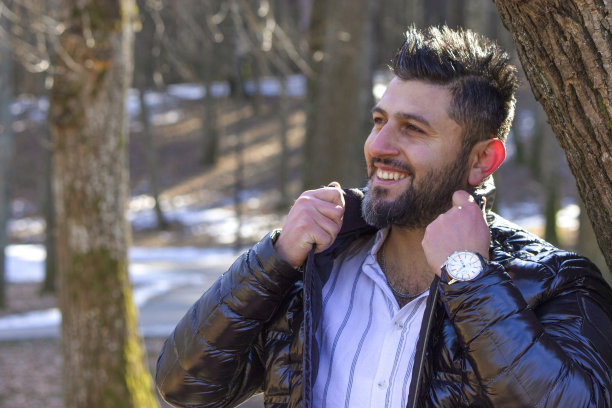 快乐微笑的土耳其男子固定他的黑色夹克，在寒冷的季节(秋、冬、早春)在公园户外，穿着时髦的衣服，自信，手表，现代发型，水平平面图片下载