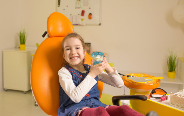 一个小女孩坐在牙科诊所的椅子上微笑着，手里拿着一个下巴模型。牙科保健，口腔卫生。图片下载