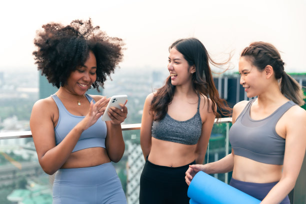 一群快乐的健身朋友在城市户外锻炼。微笑的年轻亚洲女人看着非洲女人在曼谷屋顶上玩手机。图片下载