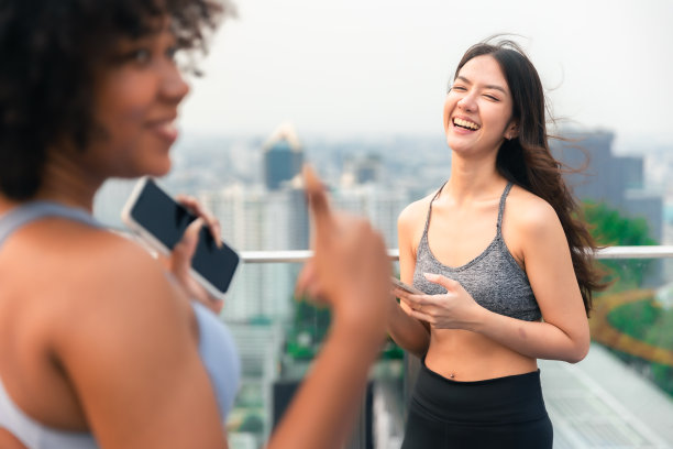 一群快乐的健身朋友在城市户外锻炼。微笑的年轻亚洲女人看着非洲女人在曼谷屋顶上玩手机。图片下载