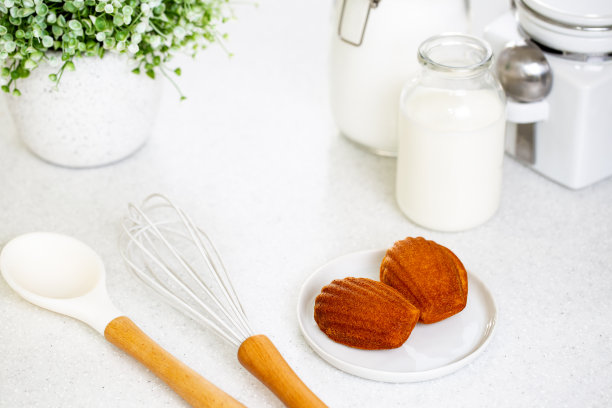 马德琳饼干或法式海绵蛋糕放在白色的盘子里，旁边放着白色的厨房用具，白底上放着糖、牛奶和面粉罐。图片下载
