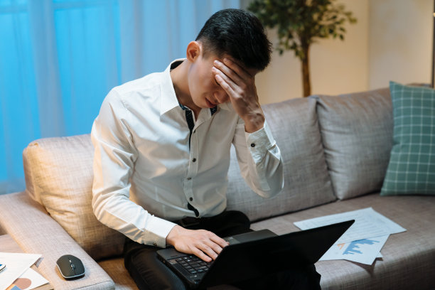 疲惫的亚洲日本人晚上在家里的客厅工作到很晚。年轻的工作人员坐在沙发上，旁边有笔记本电脑和文件图表。病男经理晚上头痛图片下载