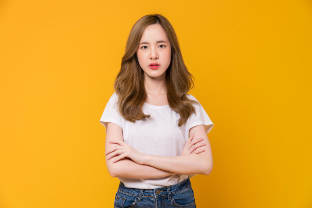 一张愤怒的年轻亚洲女孩的脸，站在橙色的背景上，双臂交叉。图片下载
