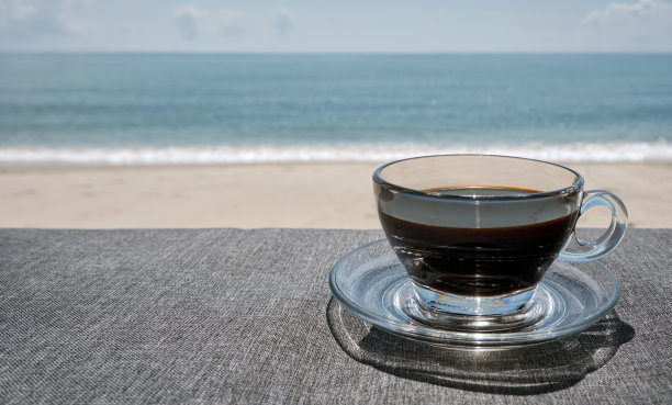 海滩和大海前的桌子上放着的透明黑咖啡杯图片下载