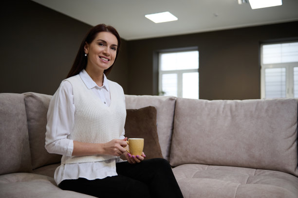 快乐迷人的女人端着一杯咖啡坐在舒适的沙发上，微笑着看着镜头。图片下载