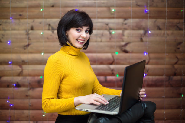 一个快乐的黑发女人在办公室里，手里拿着一台笔记本电脑，背景是一面带着花环的木墙图片下载