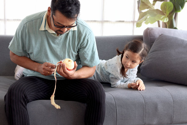 年轻帅气的爸爸为生病的女儿削苹果，可爱可爱的小女孩坐在客厅的沙发上生病，父亲照顾感冒的孩子，父亲节快乐。图片下载