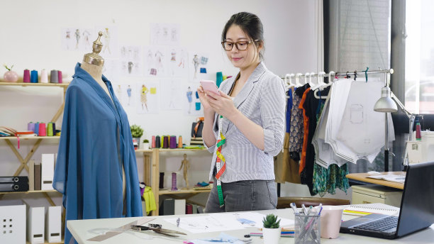 快乐的时装设计师发送照片与新的服装收集设计网上商店出售。年轻的女裁缝用手机工作在工作室车间的服装模型上图片下载