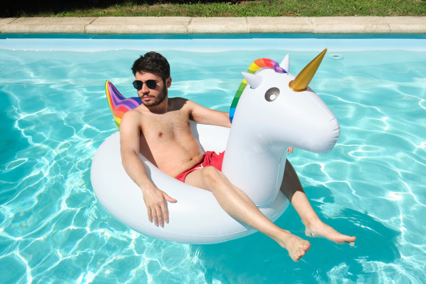 一个年轻人戴着太阳镜，戴在一个巨大的独角兽充气环上。图片下载
