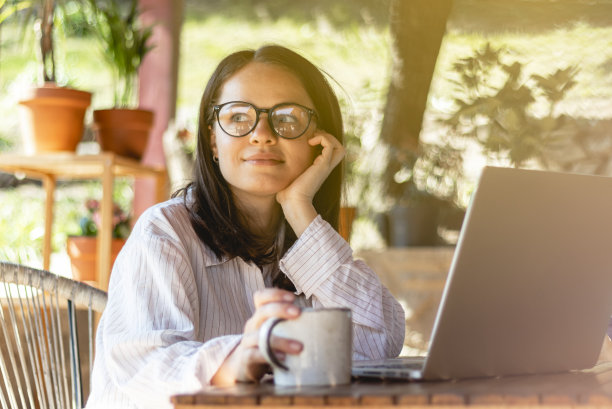 美丽微笑的年轻女子戴着眼镜，坐在桌子旁边，拿着笔记本电脑和咖啡杯。图片下载