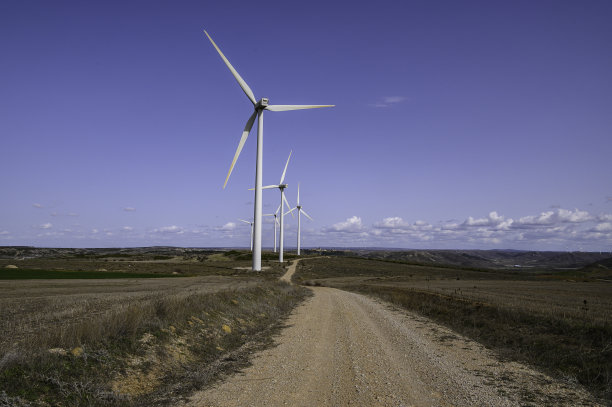 带有大叶片的白色风车能产生清洁、绿色、生态的电能。在一条迷失在无尽深处的土路边缘。背景是光天化日之下的蓝天，地平线上有小朵白云。图片下载