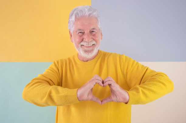 微笑的老年高加索男子与胡子制作心形的手。英俊的老祖父站在一个多彩的孤立背景表达爱和感觉图片下载