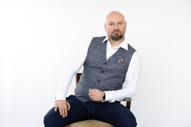 秃顶中年商人的肖像，穿着灰色背心，蓝色牛仔裤，白色衬衫坐着，肘部靠在木椅上。图片下载