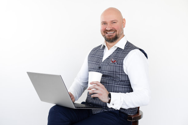 肖像快乐中年男子灰色背心，蓝色牛仔裤，白色衬衫，坐着拿着笔记本电脑和外卖咖啡。图片下载