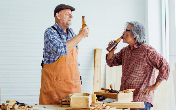 两位快乐的年长男木工穿着格子衬衫，下班后喝着啤酒，碰杯庆祝，成功制作了DIY木制家具。爱好,退休的概念。图片下载