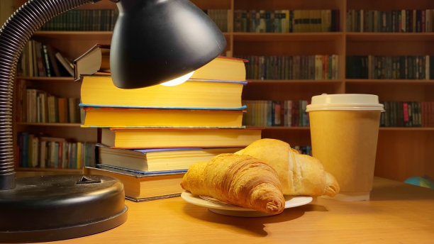 书籍、咖啡、羊角面包和旋转的台灯在图书馆的书桌上，在黑暗的房间的书架图片下载