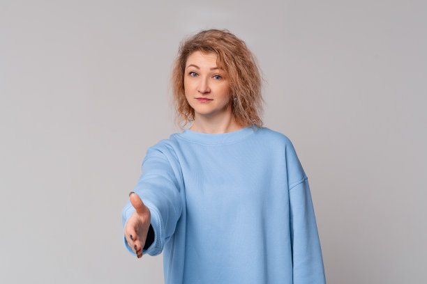 现代成熟的女人与卷曲的金发提供握手作为问候和欢迎，站在时尚的蓝色运动衫浅灰色的背景图片下载