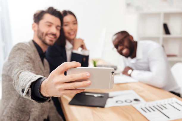 跨国商人和女商人坐在桌子旁，用手机正面相机拍照，而在办公室一起工作，隔离图片下载