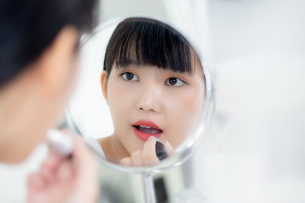 美丽的年轻亚洲女性涂口红在嘴上红润，美丽的女孩看着镜子化妆化妆品与嘴唇优雅，化妆与性感的女性，皮肤护理和健康的概念。图片下载