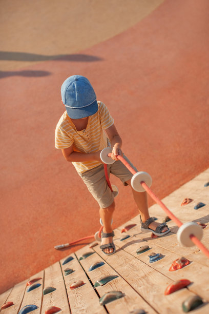 一个身穿t恤、短裤、头戴蓝色帽子、皮肤晒得黝黑的小男孩爬了上来，在一个夏日的儿童运动场上，抓着一根绳子，在攀岩图片下载