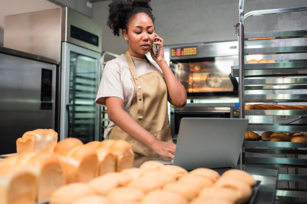 非裔美国妇女面包师看着镜头…面包师穿着厨师服，戴着厨师帽，在厨房里一起做饭。她从厨房的现代电炉中取出新鲜出炉的饼干。图片下载