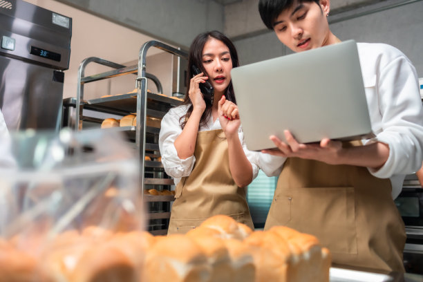 年轻的亚洲男性和非洲女性穿着围裙，在工作地点用笔记本电脑和客户打电话。情侣卖家在网上浏览手机。经营理念。零售行业。图片下载