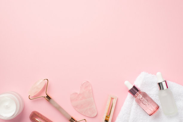 护肤美容程序概念。顶视图照片的玫瑰石英辊挂沙发夹奶油坛子两个玻璃滴管瓶和白色毛巾在粉彩粉红色的背景与空白图片下载