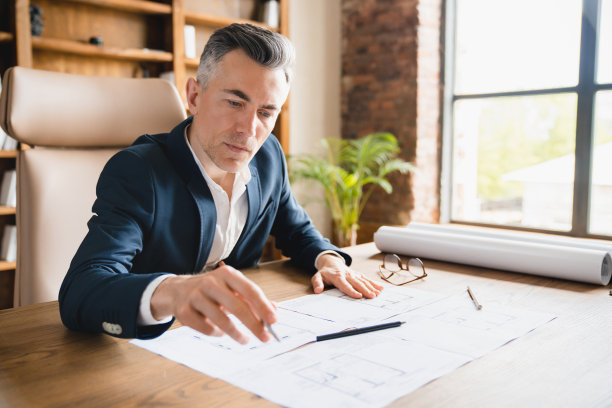 专注严肃的男性成熟中年建筑师工程师绘制蓝图，坐在办公室的办公桌前研究建筑方案，构思建筑行业。图片下载