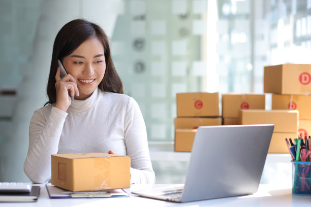 年轻的亚洲女卖家快乐的工作在笔记本电脑上使用手机打电话聊天在家里办公室书桌使用电脑在中小企业网上商店社会电子商务销售页面服务网页支持。图片下载