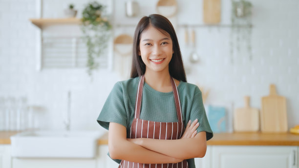 快乐的年轻亚洲女人穿着围裙，站在厨房房间。美丽的女性微笑着看着镜头，感觉很自信素材图片