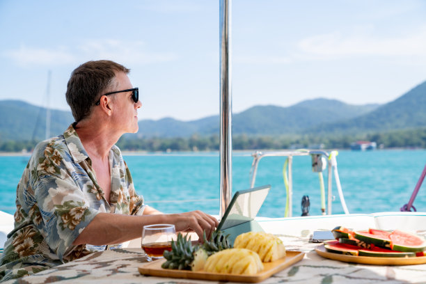 白人男子工作在数字平板电脑，而双体船航行在暑假。图片下载