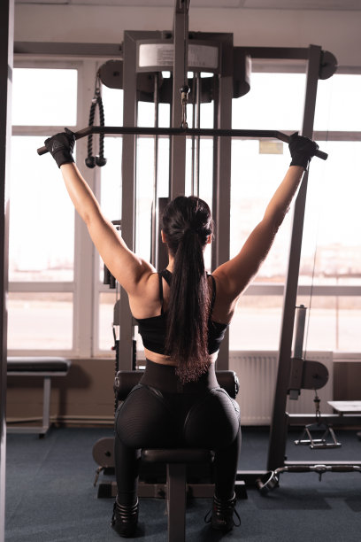 后方的年轻健身女士在黑色运动服坐在健身房机器和做手臂锻炼与重量。一个肌肉发达的女人正在锻炼。运动与健身的概念。图片下载